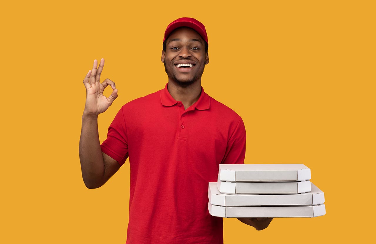 Pizzabote in rotem Shirt macht OK-Zeichen mit der Hand und hat in der anderen Pizza-Kartons